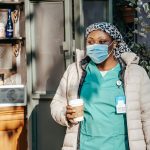 Adult black woman in nurse uniform with jacket and protective mask with headwear in city street with cup of drink near counte