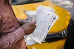 From above side view of crop anonymous ethnic male cab driver with coffee to go and newspaper talking to coworker on street —